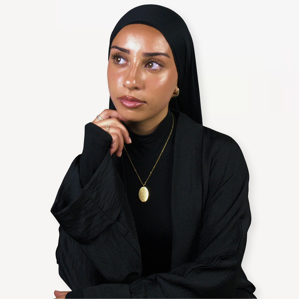 Woman wearing a black hijab and black outfit with a gold ayatul kursi necklace on a white background